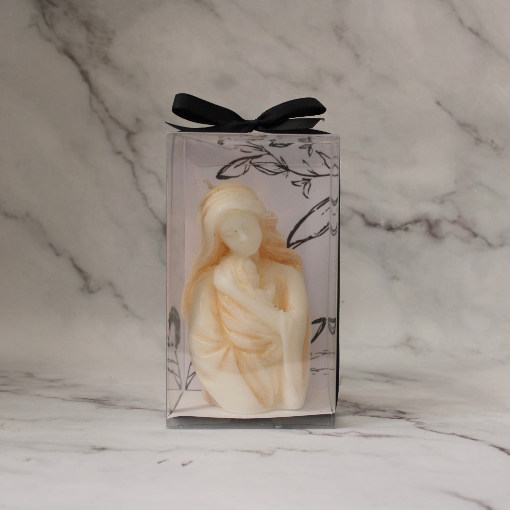 Vela artesanal en forma de Virgen con niño, presentada en caja transparente con listón negro sobre fondo de mármol.