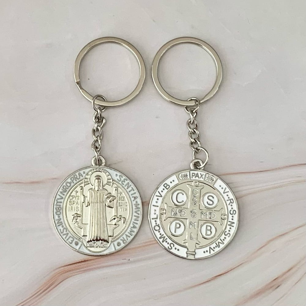 Dos llaveros metálicos plateados con la medalla de San Benito y detalles en esmalte blanco, mostrando el anverso y el reverso de la medalla sobre un fondo de mármol claro.