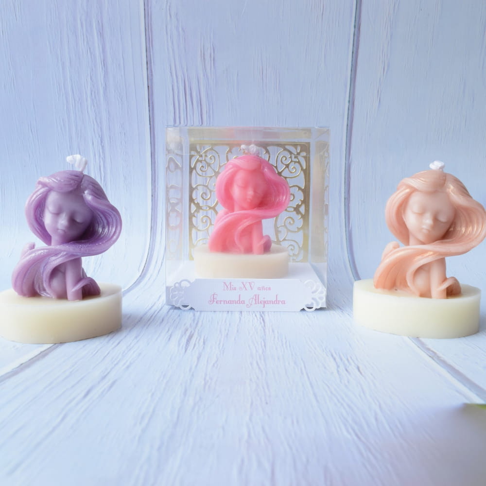Tres velas artesanales en forma de busto de joven con cabello largo en colores lila, rosa y durazno. La vela central está presentada dentro de una caja transparente con una etiqueta personalizada de quince años.