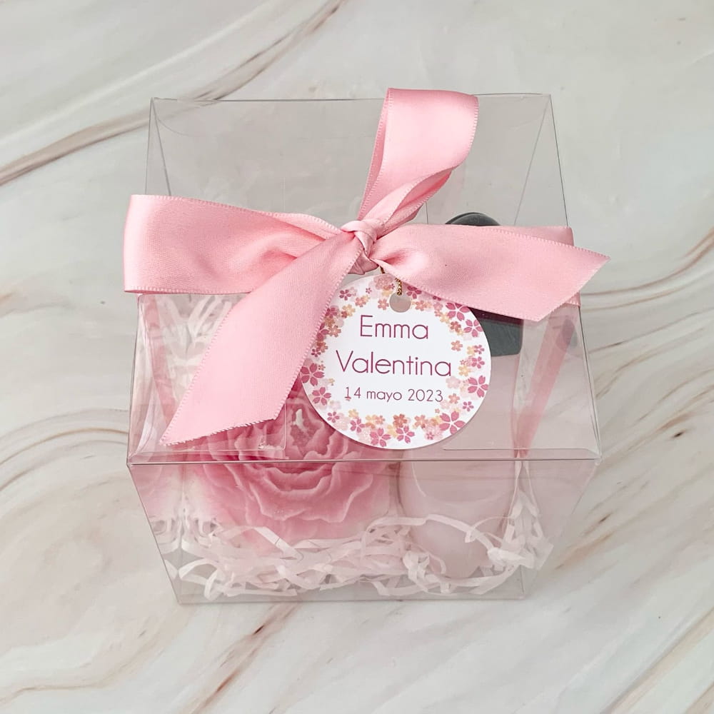 Caja transparente de recuerdo con moño de listón rosa y etiqueta personalizada, que contiene una vela artesanal en forma de flor rosa y un perfume en forma de zapatilla.
