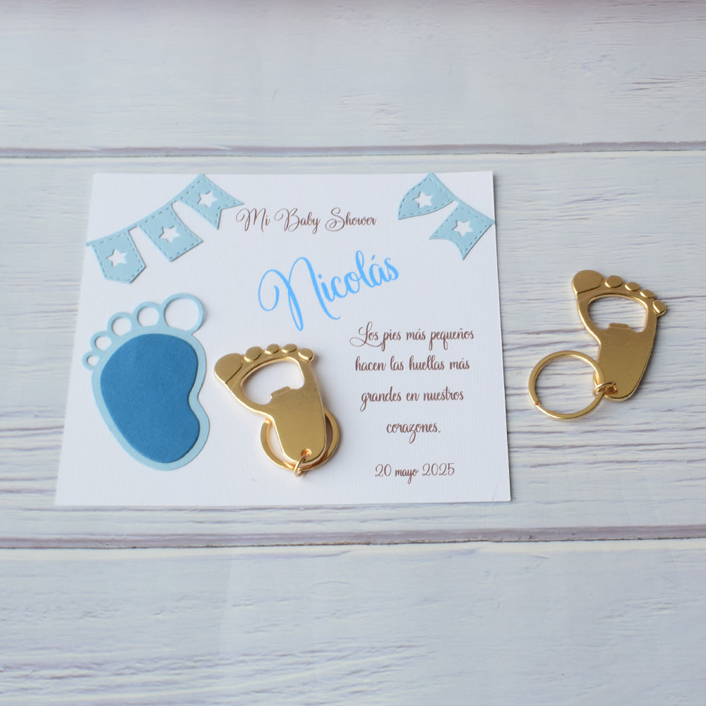Recuerdo para baby shower con llaveros destapadores en forma de pie y tarjeta personalizada sobre madera blanca.