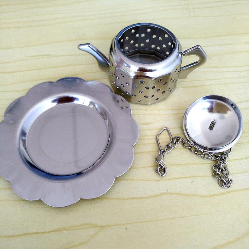 Infusor de té metálico en forma de pequeña tetera con plato y cadena, ideal como recuerdo para eventos, sobre superficie de madera clara.