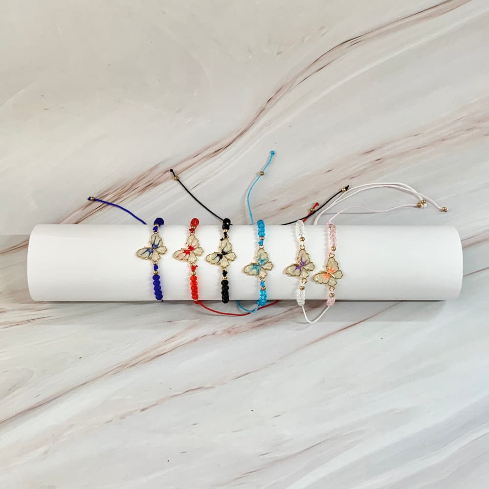 Seis pulseras tejidas ajustables en diferentes colores (azul oscuro, rojo, negro, celeste, blanco y rosa), decoradas con cuentas y un dije central de mariposa esmaltada con bordes dorados, exhibidas sobre un cilindro blanco con fondo de mármol.