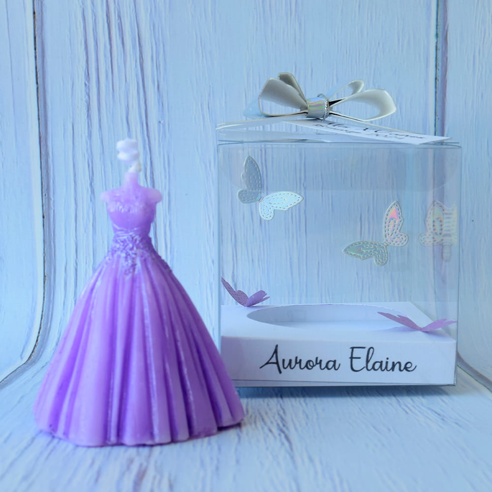 Vela artesanal color lila en forma de vestido de quinceañera, junto a una caja transparente decorada con mariposas plateadas y base personalizada.