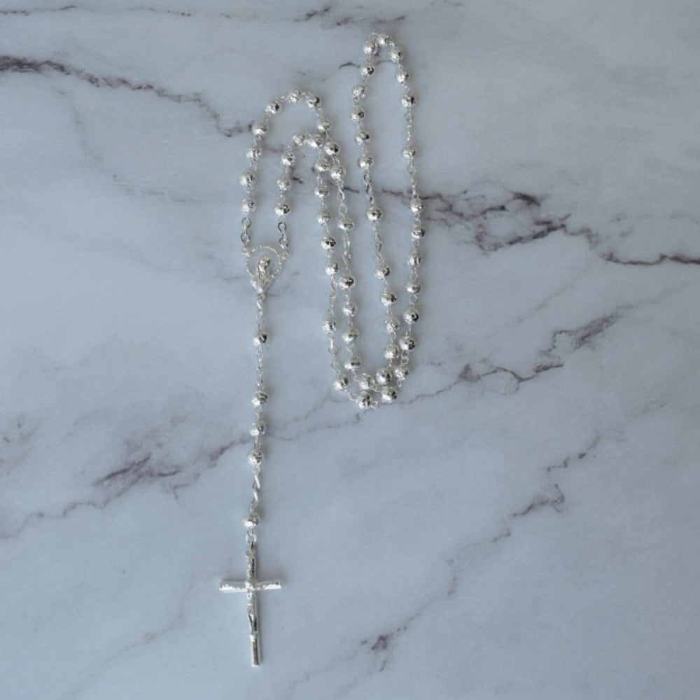 Rosario plateado con cuentas esféricas y cruz de diseño clásico sobre fondo de mármol, ideal para recuerdos religiosos.