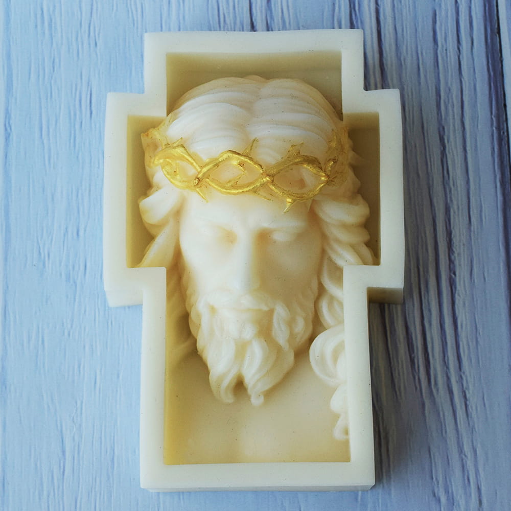 Vela artesanal blanca en forma de cruz con el rostro de Jesucristo en relieve y una corona de espinas pintada de dorado.