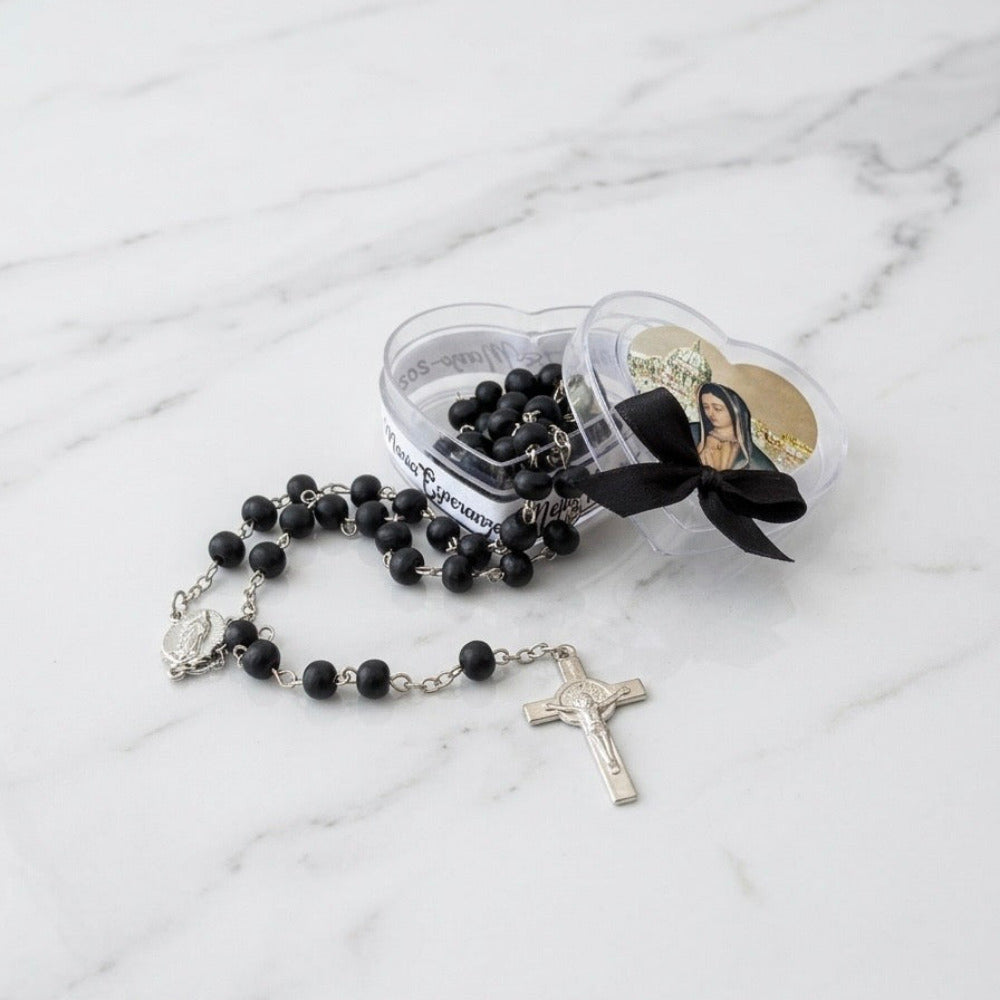Rosario de cuentas negras con cruz de metal, presentado en una caja transparente en forma de corazón con moño negro y estampa de la Virgen, ideal como recuerdo para aniversario luctuoso.